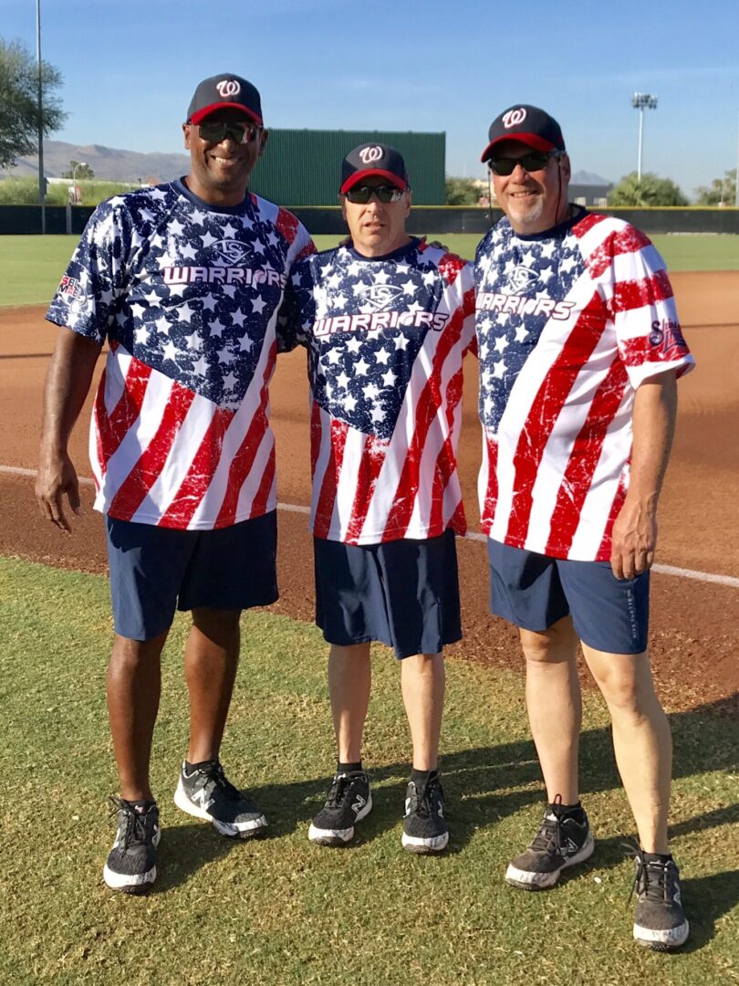 Three men standing on a baseball field wearing patriotic shirts.